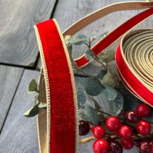 May include: Close-up of a red velvet ribbon with a gold trim. The ribbon is partially unrolled, revealing a roll of the same ribbon. Red berries and green leaves are in the background. The ribbon is likely used for crafts or holiday decorations.