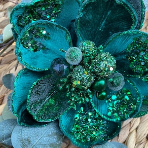 Op de afbeelding: Close-up van een grote, teal-kleurige bloemdecoratie met glitteraccenten. De bloem heeft meerdere lagen bloemblaadjes en een cluster van kralenversieringen in het midden. De achtergrond is een geweven textuur.