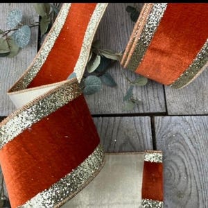 Pode incluir: Uma fita decorativa larga com uma faixa central de veludo laranja queimado, ladeada por duas bordas de glitter dourado brilhante. A fita está disposta em laços soltos, exibindo o seu tecido texturizado e o brilho metálico.