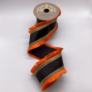 Pode incluir: Um carretel de fita preta com franjas laranja e acabamento dourado. A fita é rotulada "d.stevens fine ribbons". A fita é desenrolada, mostrando o tecido preto com franjas laranja e acabamento dourado.