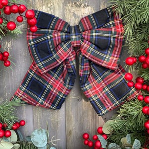 May include: A large decorative bow featuring a classic black, red, green, and yellow plaid tartan pattern. The bow is positioned centrally, surrounded by artificial evergreen pine branches and clusters of small, bright red berries.