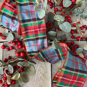May include: A wired ribbon featuring a classic tartan pattern in shades of red, green, blue, and yellow with a solid red stitched edge. The ribbon is draped over a decorative arrangement of artificial eucalyptus leaves and small red berries.