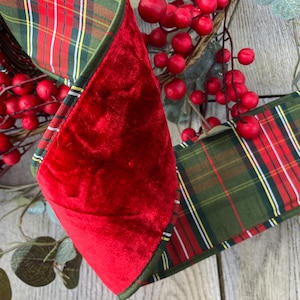 May include: A decorative ribbon featuring a dual-sided design with one side in solid red velvet and the other side in a green, red, white, and yellow plaid pattern. The ribbon is displayed alongside artificial red berries and green foliage.