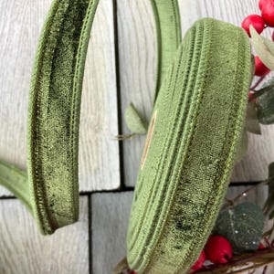 May include: A spool of olive green velvet ribbon with a textured surface and stitched edges. The ribbon is wound into a circular coil, with a loose end extending outward, showcasing the soft, plush fabric and consistent deep green color.