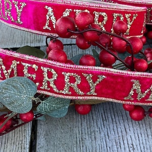 May include: A bright pink velvet ribbon featuring the repeating gold glitter text MERRY. The ribbon is draped over a decorative branch with small red berries and muted green leaves, creating a layered and textured appearance.