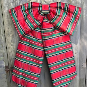 May include: A large decorative bow made of fabric featuring a red, green, and yellow plaid pattern. The bow has two wide, long tails hanging downwards and a central knot, with a structured, stiff appearance and a subtle sheen on the material.
