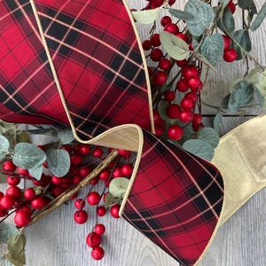 May include: A wide wired ribbon featuring a red and black plaid pattern with thin gold metallic lines. The ribbon has a gold metallic edge and is displayed alongside a decorative branch with red berries and green leaves on a light wood surface.