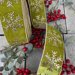May include: A roll of olive green velvet ribbon featuring a repeating pattern of gold glitter snowflakes. The ribbon has finished edges and is displayed alongside sprigs of artificial red berries and green eucalyptus leaves on a wooden surface.