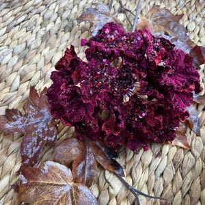 May include: A close-up of a decorative floral arrangement featuring a cluster of deep burgundy flowers with a textured appearance. The flowers are adorned with glitter and sit atop dried, brown leaves. The background is a woven, light brown surface.