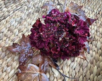 May include: A close-up of a decorative floral arrangement featuring a cluster of deep burgundy flowers with a textured appearance. The flowers are adorned with glitter and sit atop dried, brown leaves. The background is a woven, light brown surface.