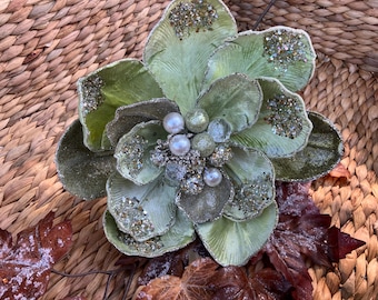 May include: A large, decorative flower with layered, sage green petals dusted with glitter. The center features pearl and silver bead accents. The flower rests on a woven background with frosted brown leaves, suggesting a fall or winter theme.