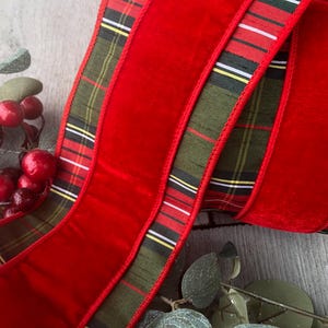 May include: Close-up of a red velvet ribbon with a green and plaid border. The ribbon is curled and layered, with a festive appearance. Red berries and green leaves are in the background, suggesting a holiday theme.