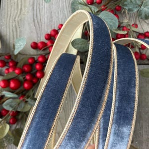 May include: A length of navy blue velvet ribbon featuring a soft, textured pile surface and finished with a metallic gold stitched edge along both sides. The ribbon is coiled in loose loops, displaying its deep blue colour and gold trim.