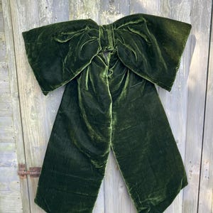 May include: A large, dark forest green velvet bow featuring two wide, flowing tails and a central knot. The fabric has a soft, light-reflecting texture with visible folds and creases throughout the structure, creating a voluminous appearance.