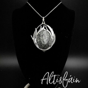 May include: A silver necklace with a large, round pendant featuring a gray and white stone set in a silver frame. The pendant is surrounded by a silver design that resembles a stylized flame. The necklace is on a black background with the word "Altisfain" written in white.
