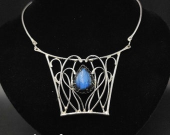 Puede incluir: Un collar de plata con un diseño delicado e intrincado que presenta una piedra preciosa azul en forma de lágrima. El collar tiene un estilo de gargantilla y está adornado con un patrón geométrico.