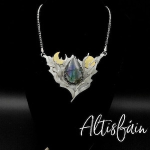 May include: A silver and gold necklace with a large iridescent gemstone pendant. The pendant is surrounded by a silver design that resembles flames and crescent moons. The necklace is on a black background with the word "Altisfain" written in white.
