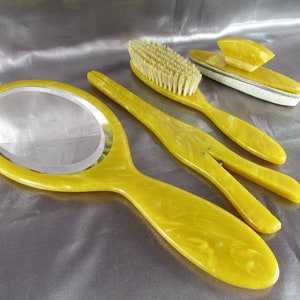 May include: A vintage yellow vanity set with a hand mirror, a hairbrush, and a comb. The set is made of marbled plastic.