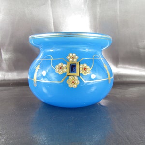 Puede incluir: Un pequeño jarrón de vidrio azul con detalles pintados en oro y blanco. El jarrón tiene un cuerpo redondo y una boca ancha. Está decorado con una cruz dorada y diseños florales.