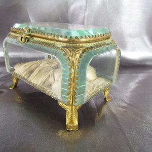 May include: A small, rectangular glass trinket box with gold-toned metal accents and feet. The box has a clear glass lid and a white fabric lining.
