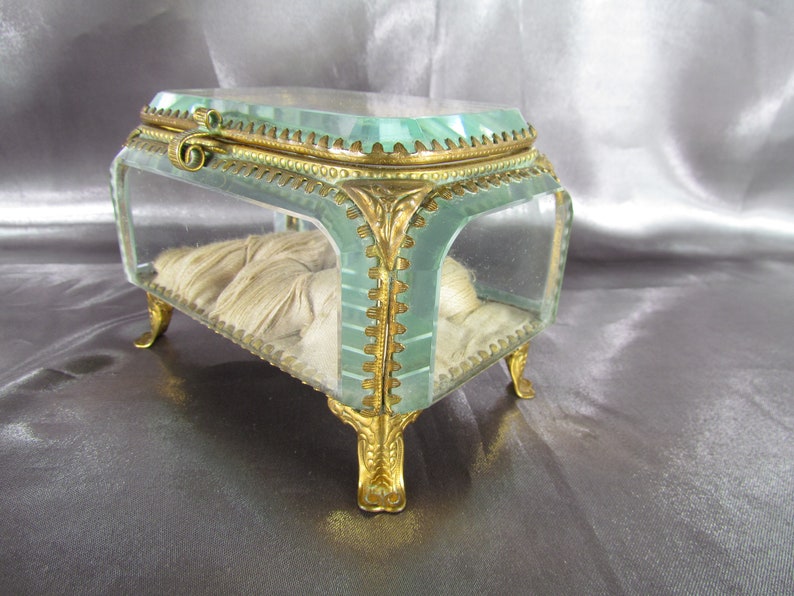 May include: A small, rectangular glass trinket box with gold-toned metal accents and feet. The box has a clear glass lid and a white fabric lining.