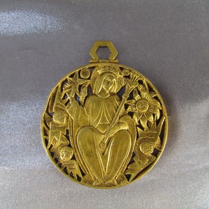 May include: A gold-colored pendant with a detailed relief design. The pendant features a central figure holding a staff, surrounded by floral and star motifs. The pendant has a hexagonal loop at the top for hanging.