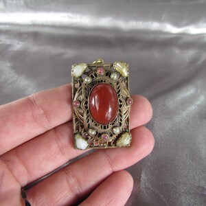 May include: A gold-toned pendant with a red carnelian stone surrounded by white and pink stones. The pendant is in a square shape with intricate filigree details.