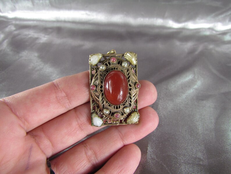May include: A gold-toned pendant with a red carnelian stone surrounded by white and pink stones. The pendant is in a square shape with intricate filigree details.