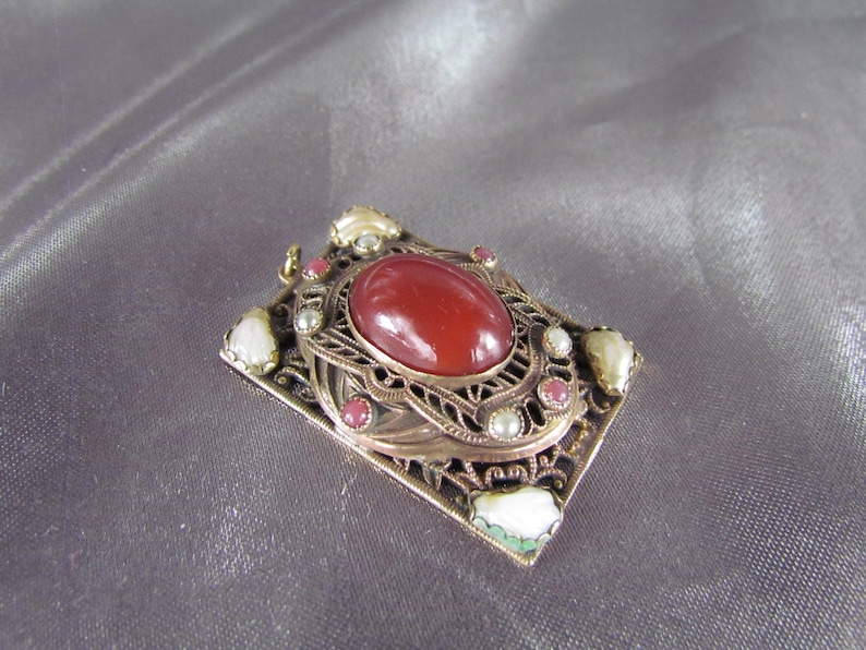 May include: A gold-toned pendant with a red carnelian stone set in a filigree frame. The frame is decorated with white pearls and pink and green stones.