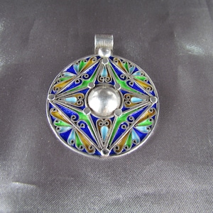 May include: A silver pendant with a circular design. The pendant features a central silver sphere surrounded by a colorful enamel pattern in blue, green, yellow, and light blue. The pendant has a silver loop at the top for a chain.