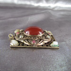 May include: A gold-toned pendant with a red carnelian stone surrounded by smaller white and pink stones. The pendant has a filigree design and is set in a square frame.