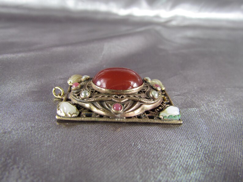 May include: A gold-toned pendant with a red carnelian stone surrounded by smaller white and pink stones. The pendant has a filigree design and is set in a square frame.