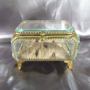 May include: A small, clear glass jewelry box with gold trim and feet. The box has a padded interior and a hinged lid.