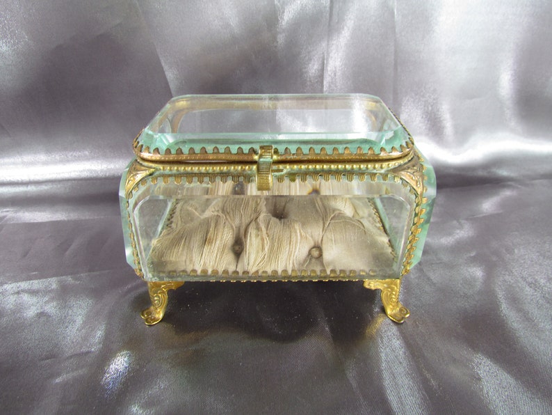 May include: A small, clear glass jewelry box with gold trim and feet. The box has a padded interior and a hinged lid.