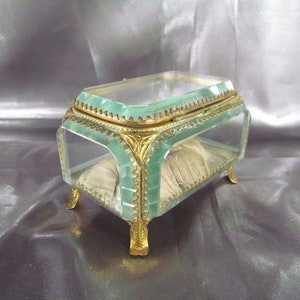 May include: A small, rectangular glass trinket box with a green tint. The box has gold-colored trim and feet. The box is open and has a white fabric lining.