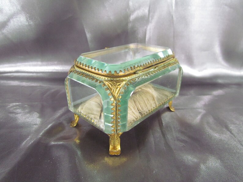May include: A small, rectangular glass trinket box with a green tint. The box has gold-colored trim and feet. The box is open and has a white fabric lining.