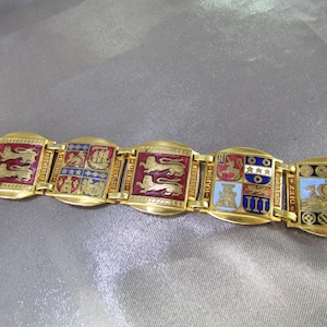 May include: A gold-tone link bracelet featuring rectangular panels with colorful enamel heraldic designs, including lions, ships, and castles. Each panel is connected by small gold-tone metal links, and the text NORMANDIE is visible on the side of the links.