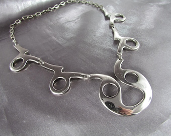 Puede incluir: Un collar de plata con un diseño único y abstracto que presenta múltiples bucles y círculos interconectados.