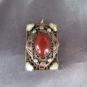 May include: A gold-tone pendant with a red carnelian stone surrounded by a filigree design. The pendant is decorated with small white pearls and pink stones.