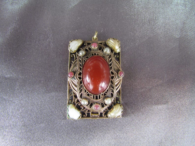 May include: A gold-tone pendant with a red carnelian stone surrounded by a filigree design. The pendant is decorated with small white pearls and pink stones.