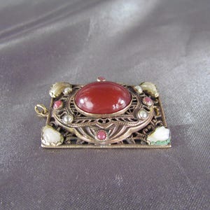 May include: A red carnelian gemstone set in a gold-toned filigree pendant with white and pink accents.