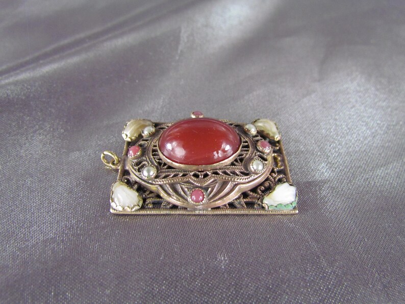 May include: A red carnelian gemstone set in a gold-toned filigree pendant with white and pink accents.