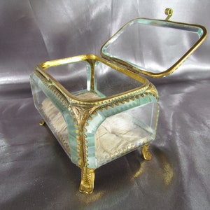 May include: A glass jewelry box with a gold metal frame and feet. The box has a hinged lid and is lined with white fabric.