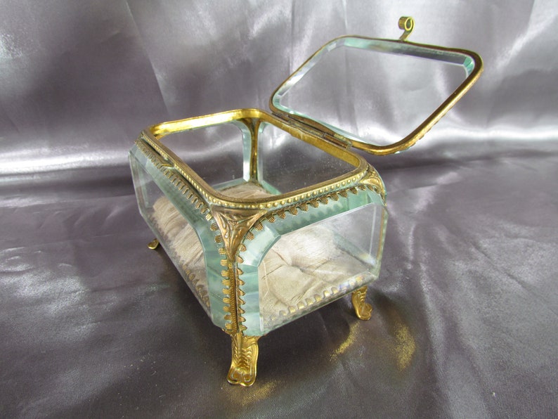 May include: A glass jewelry box with a gold metal frame and feet. The box has a hinged lid and is lined with white fabric.