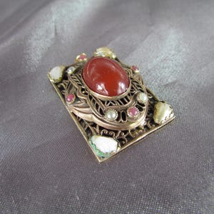 May include: A gold-toned filigree brooch with a red carnelian stone surrounded by smaller white and pink stones.