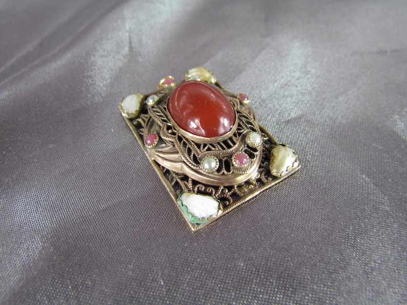 May include: A gold-toned filigree brooch with a red carnelian stone surrounded by smaller white and pink stones.