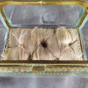 May include: A small, glass, square-shaped box with a gold-colored frame. The box has a padded interior with a tufted design.