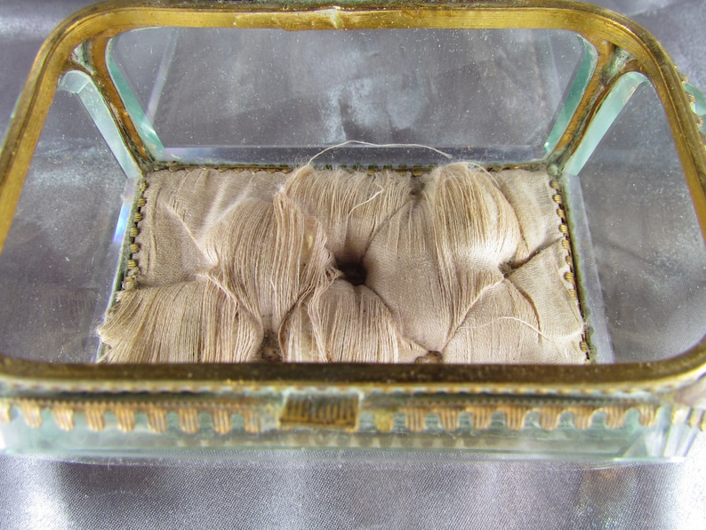 May include: A small, glass, square-shaped box with a gold-colored frame. The box has a padded interior with a tufted design.