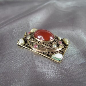 May include: A gold-toned pendant with a red carnelian stone surrounded by white and pink pearls and green stones.