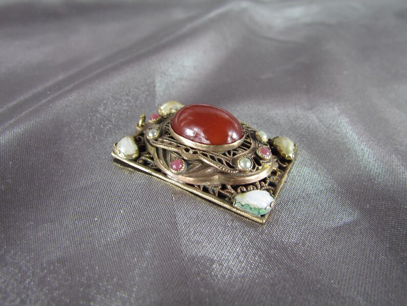 May include: A gold-toned pendant with a red carnelian stone surrounded by white and pink pearls and green stones.